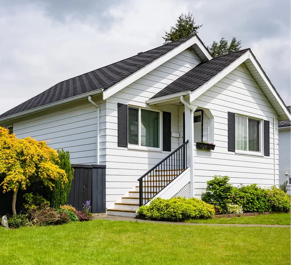 A charming white house with black shutters and a sloped roof, surrounded by lush greenery and a well-maintained lawn.