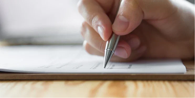 A close-up of a hand holding a silver pen, poised above a sheet of paper with printed lines, on a wooden surface.