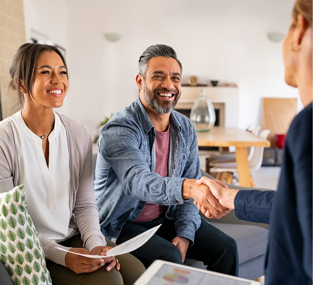 A woman and a man sit on a couch, while another woman shakes hands with the man, discussing a potential agreement.