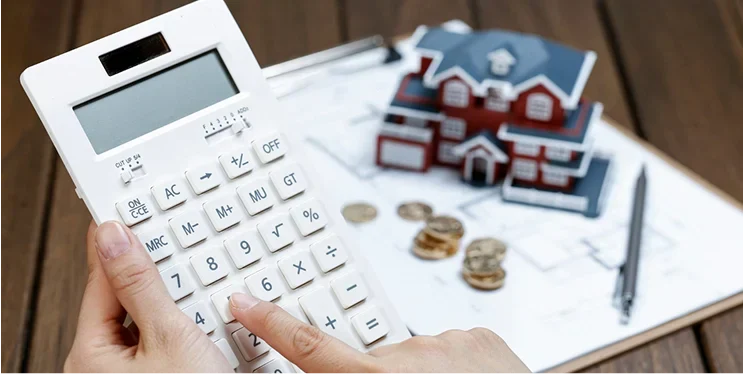 A person uses a calculator to analyze real estate finances, with a miniature house and coins on a financial document in the background.