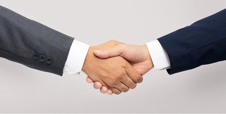 Two hands shake in a business setting, one dressed in a dark suit and the other in a light shirt, symbolizing agreement and partnership.